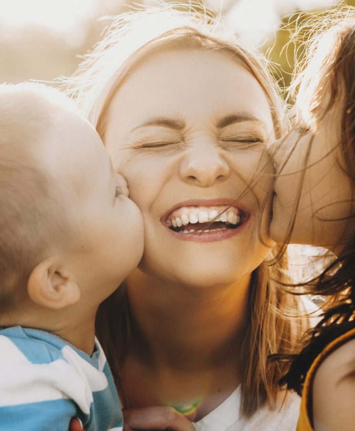 Close up portrait of lovely young mother laughing with closed eyes while her kids is kissing her on the cheeks outdoor against sunset.